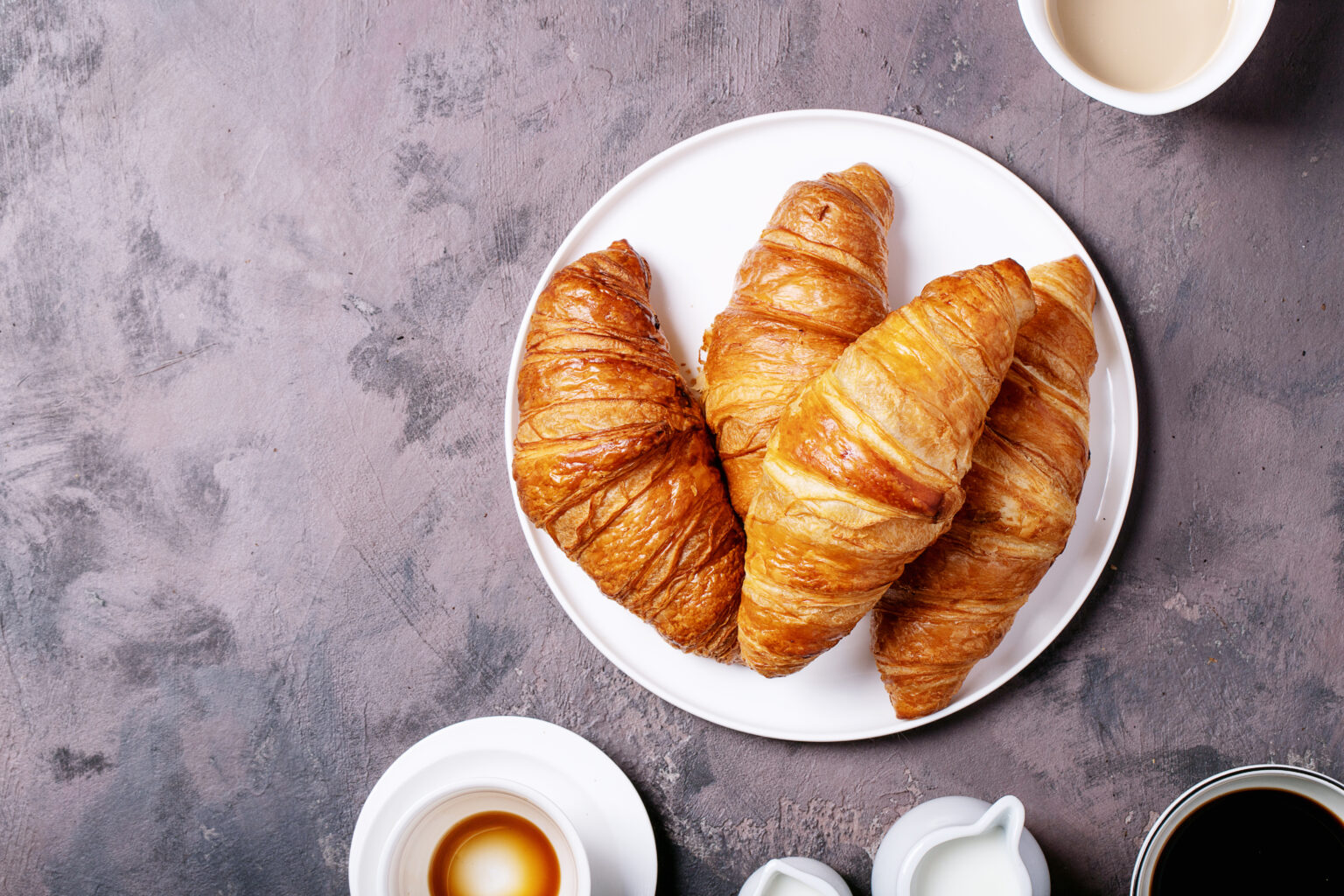 Fresh coffee in white ceramic cups served with croissants over brown texture brown background. Top view, flat lay