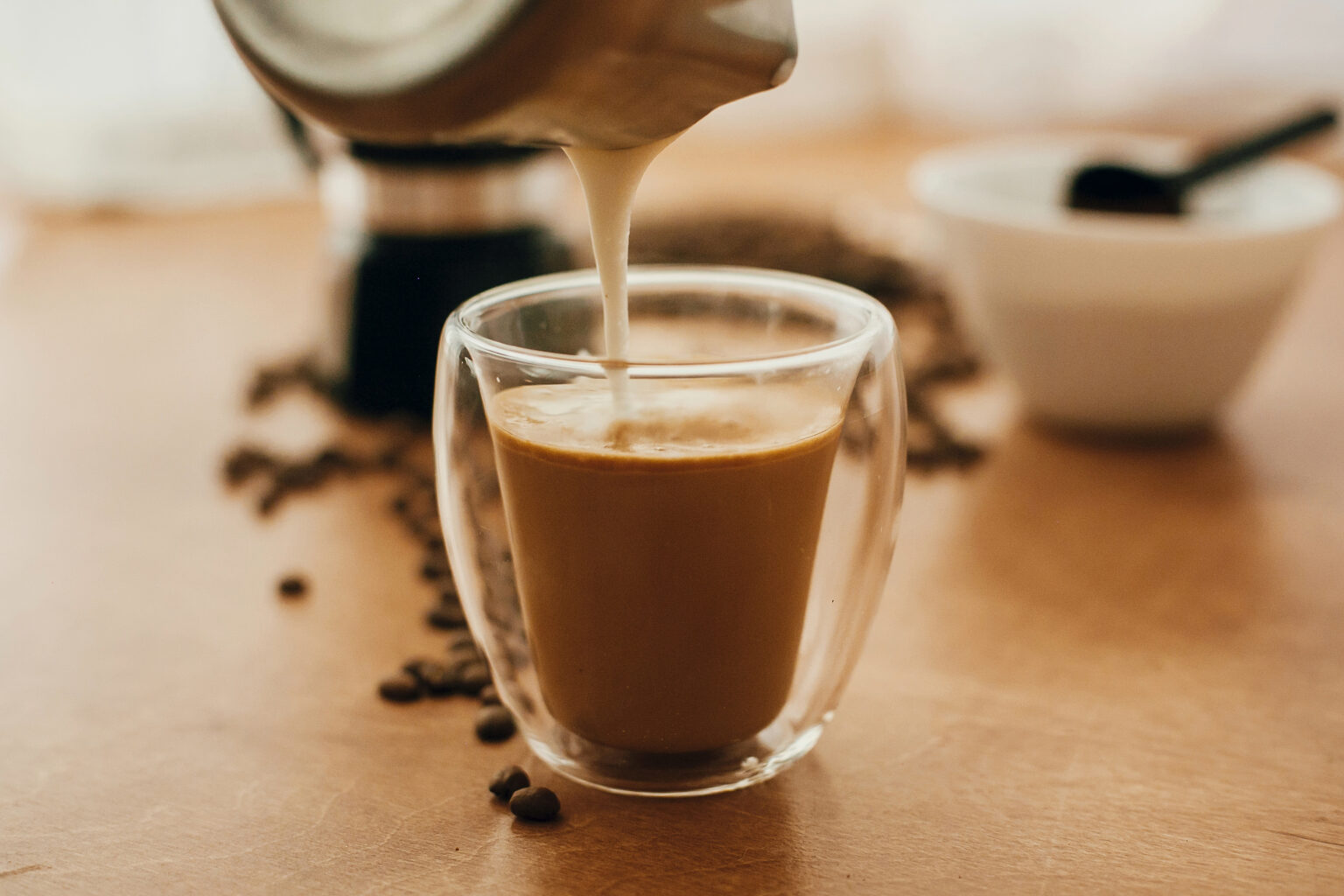 Pouring milk into coffee in glass cup on background of roasted coffee beans, grounded coffee, geyser coffee maker on wooden table. Making latte or cappuccino