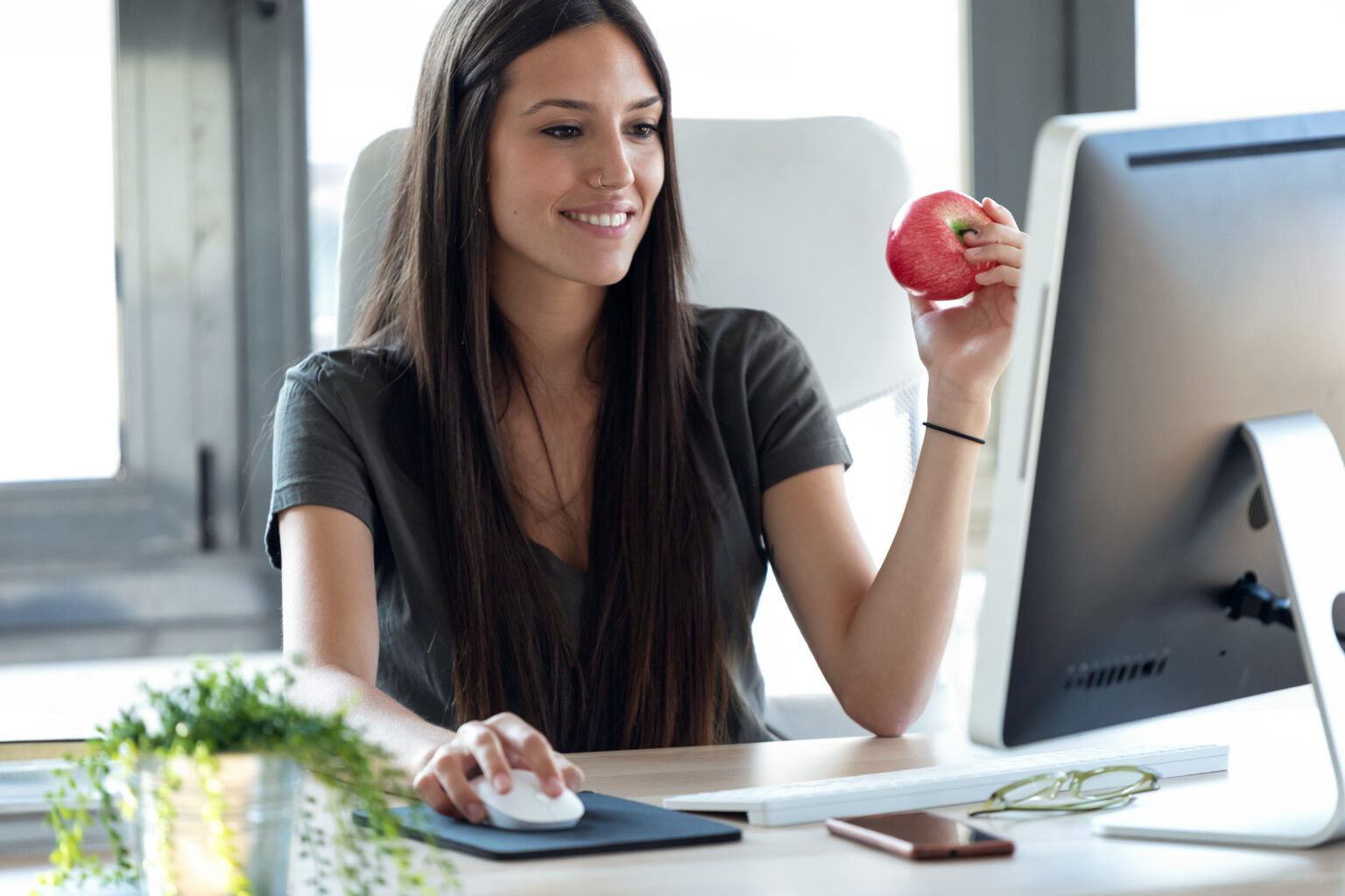 Shot of smiling young business woman eating a red apple while working with computer in the office.